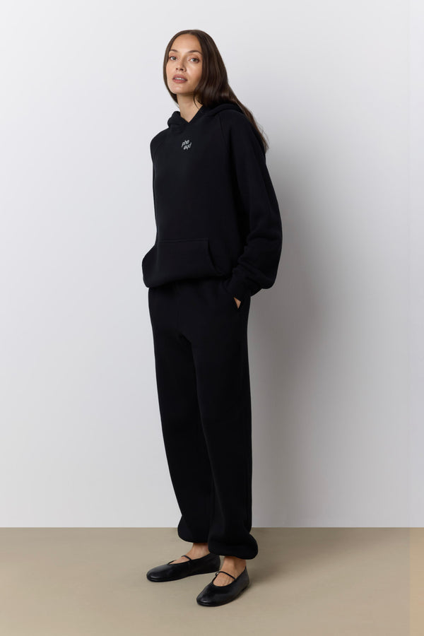 A woman modeling a black hooded sweatshirt and matching black wide-leg sweatpants against a light gray wall and beige floor. |M1