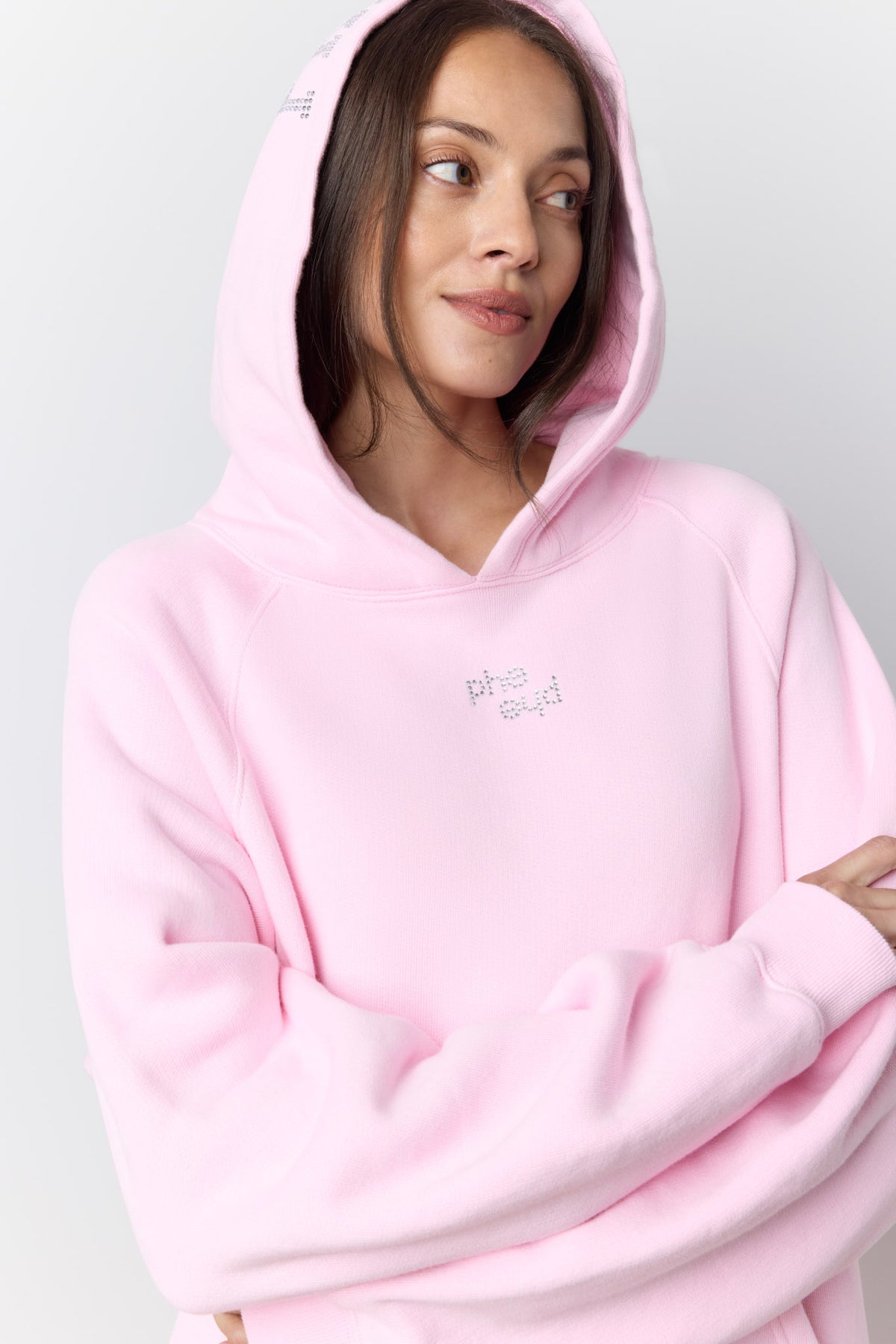 A woman wearing a light pink pullover hoodie with a small rhinestone 'phe phe' logo on the center chest, photographed against a light gray studio background.