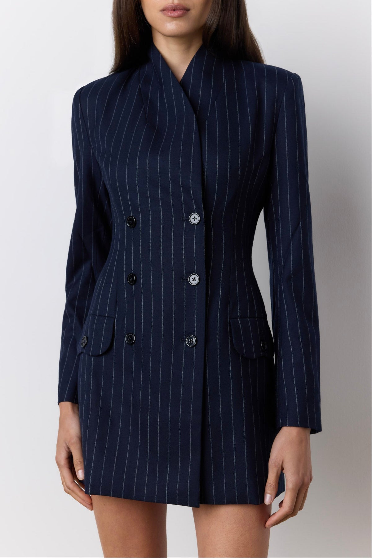 A woman wearing a navy blue double-breasted pinstripe blazer dress with buttons, shown from the head down against a light gray studio background.