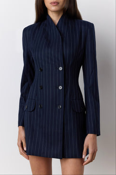 A woman wearing a navy blue double-breasted pinstripe blazer dress with buttons, shown from the head down against a light gray studio background.