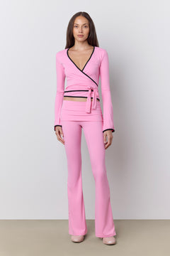 A woman modeling a bright pink two-piece outfit consisting of a long-sleeve wrap-style top with black trim and matching high-waisted flared pants, photographed against a plain light gray studio background.