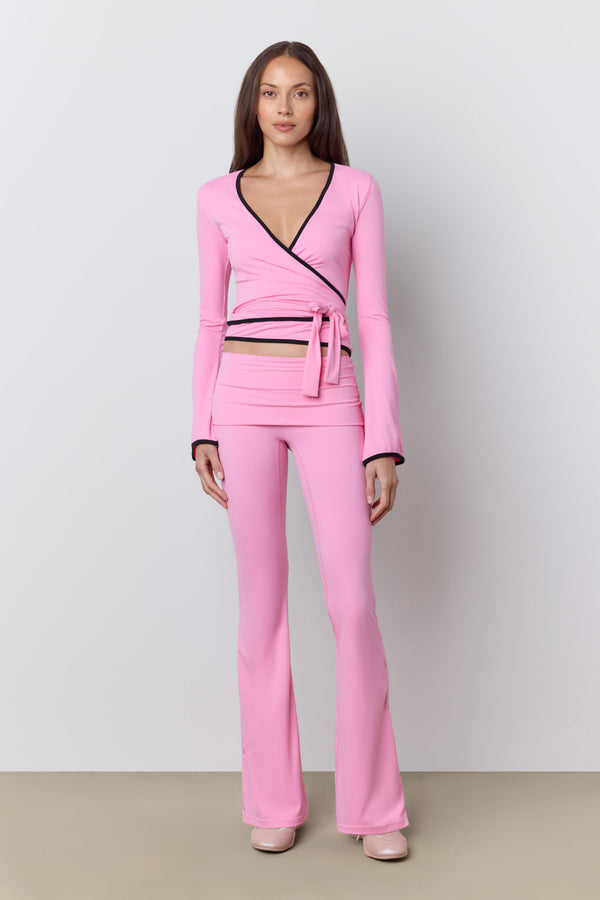 A woman modeling a bright pink two-piece outfit consisting of a long-sleeve wrap-style top with black trim and matching high-waisted flared pants, photographed against a plain light gray studio background. |M1