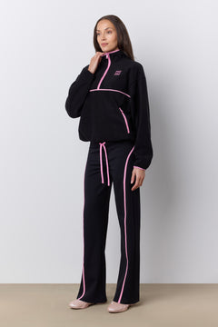 A woman modeling a black tracksuit with bright pink piping and a pink drawstring, standing against a light gray studio wall.