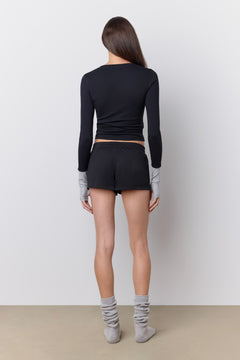 A woman stands with her back to the camera wearing a black long-sleeve athletic top, black shorts, and light gray socks against a plain light gray wall and beige floor.