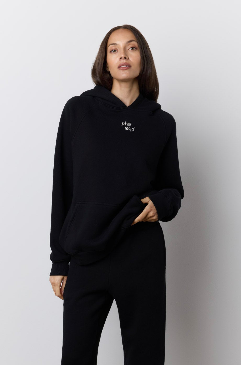 A woman modeling a black two-piece loungewear set consisting of a pullover hoodie with a small rhinestone "phe phe" logo on the chest and matching wide-leg sweatpants, set against a neutral studio background.