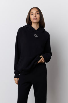 A woman modeling a black two-piece loungewear set consisting of a pullover hoodie with a small rhinestone "phe phe" logo on the chest and matching wide-leg sweatpants, set against a neutral studio background.