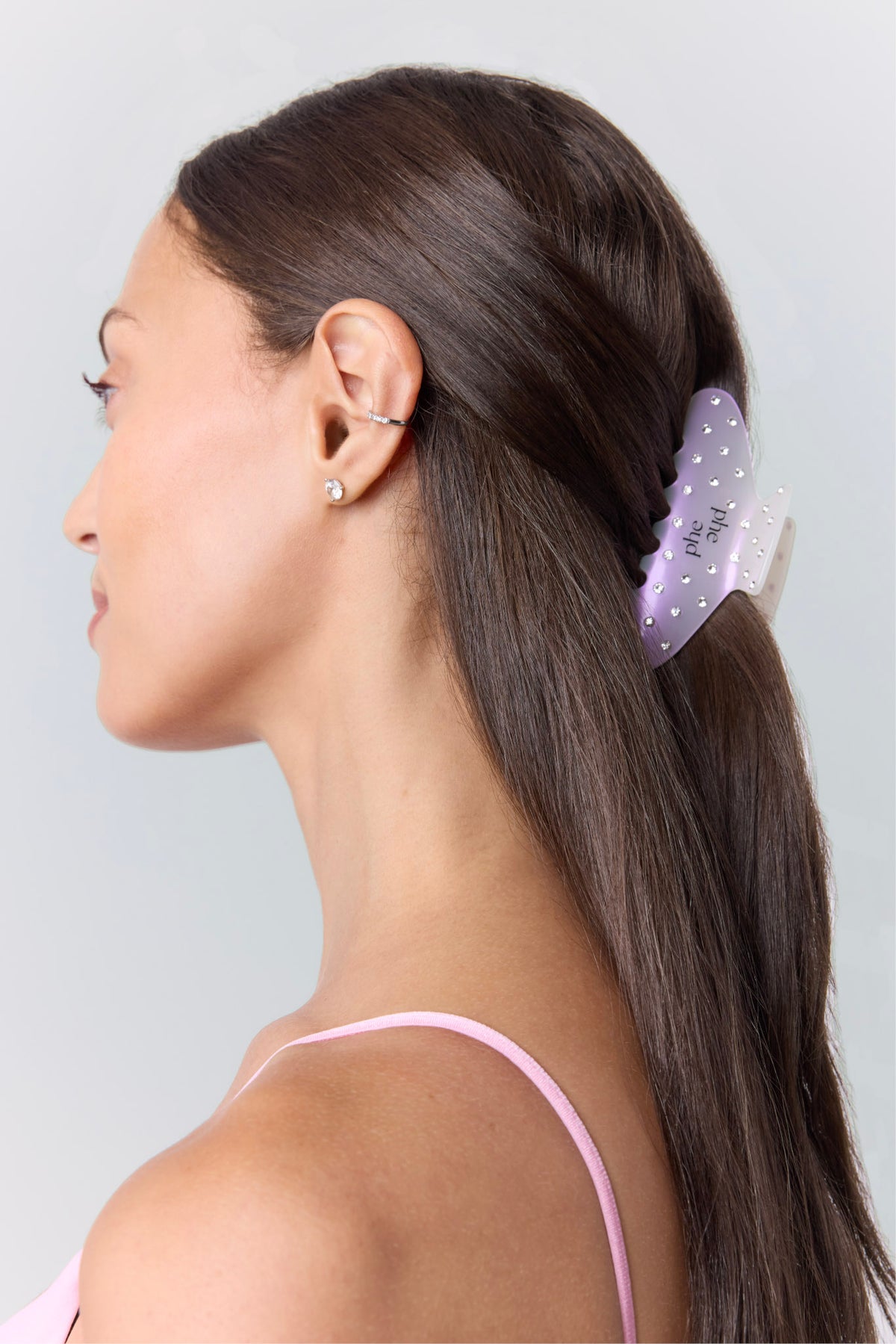 A woman shown in side profile with long dark brown hair, wearing an opal claw hair clip with silver rhinestones, and "phe phe" logo, holding her hair in place against a light gray background.