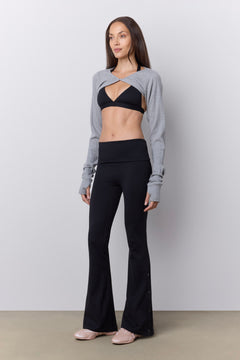 A woman stands against a light gray studio wall wearing a gray long-sleeve bolero style top over a black sports bra, paired with black flared leggings and pink sneakers.
