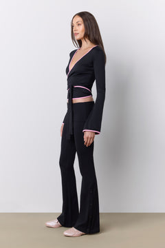 A woman modeling a black long-sleeve wrap-style top and matching flared pants set with thin pink trim, standing against a light gray studio wall.