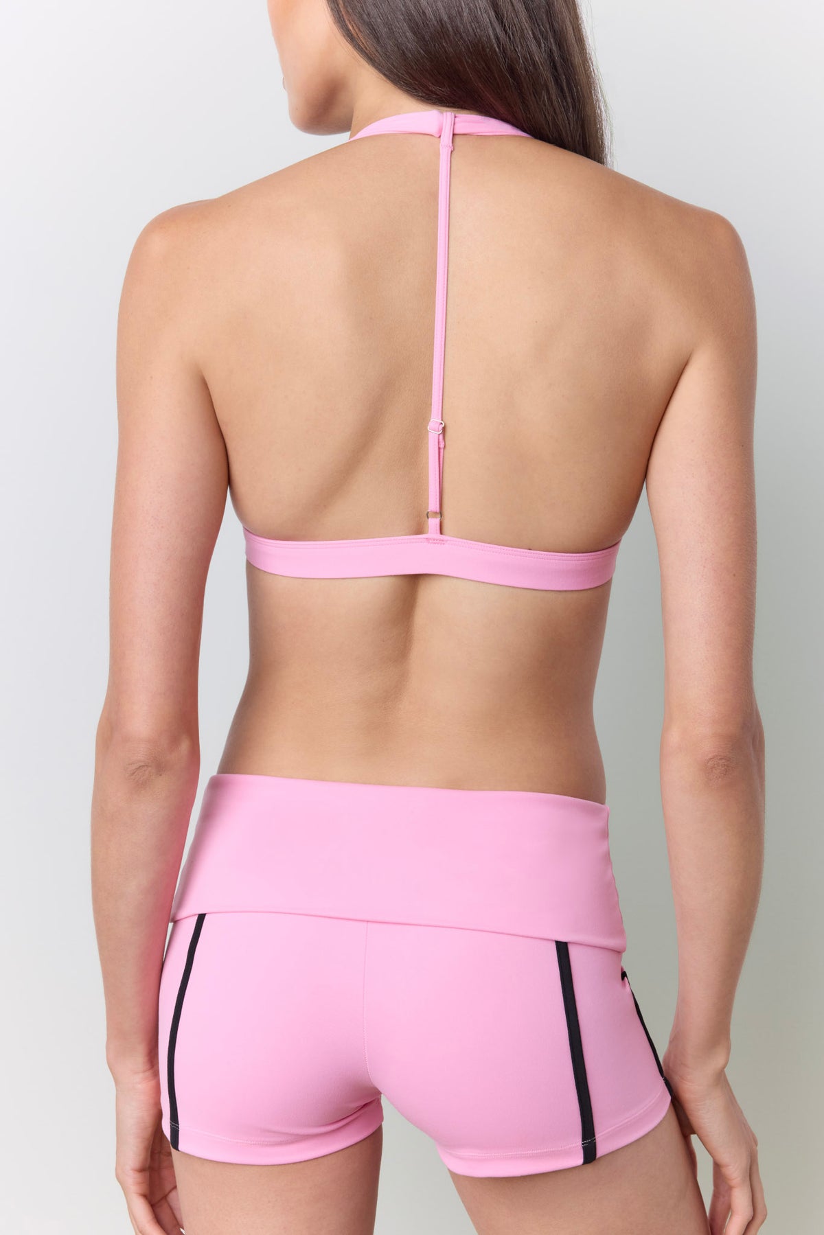 A woman shown from the back wearing a pink athletic halter sports bra and matching high-waisted shorts with black side stripes, against a light gray studio background.