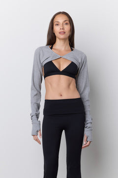 A woman stands against a light gray wall wearing a gray long-sleeve bolero style top over a black sports bra, paired with black flared leggings and pink sneakers.