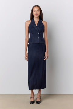 A woman stands against a light gray studio wall wearing a sleeveless navy pinstripe double-breasted vest and matching maxi skirt, paired with black Mary Jane heels.