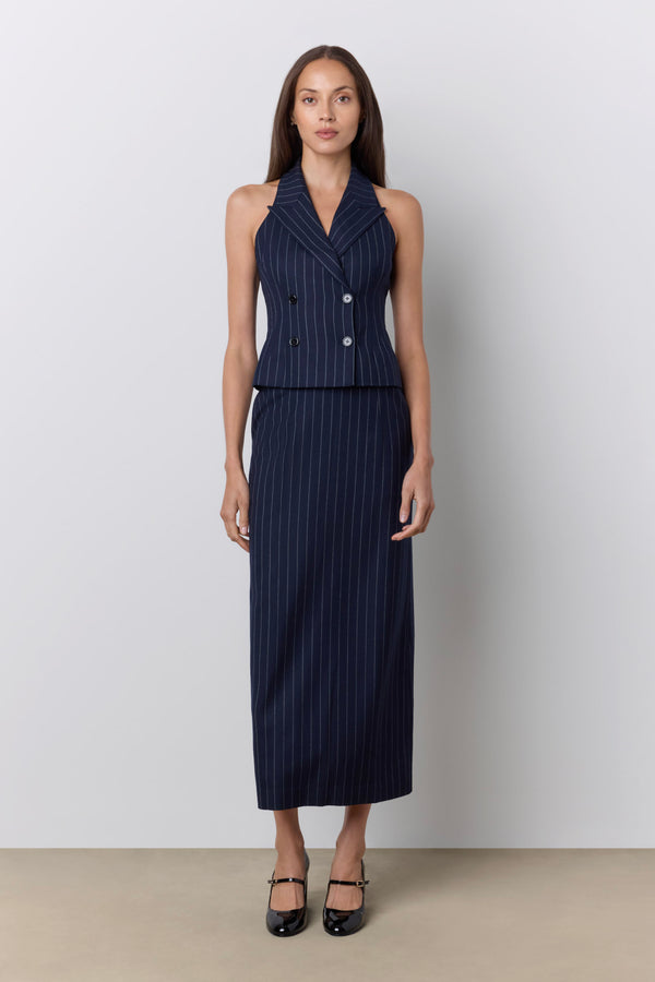 A woman stands against a light gray studio wall wearing a sleeveless navy pinstripe double-breasted vest and matching maxi skirt, paired with black Mary Jane heels. |M1