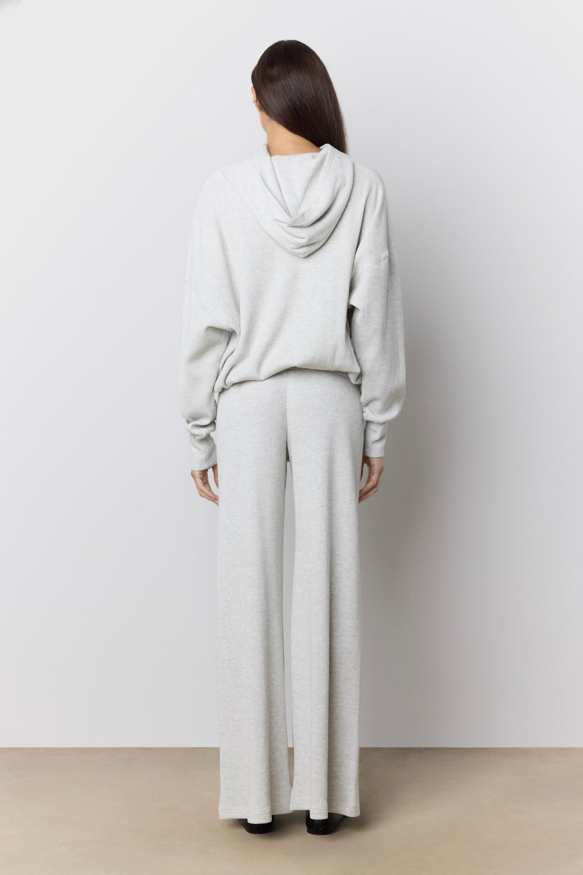 A woman viewed from behind wearing a light gray waffle knit lounge set consisting of a hooded sweatshirt and matching wide-leg pants, standing against a neutral studio wall.