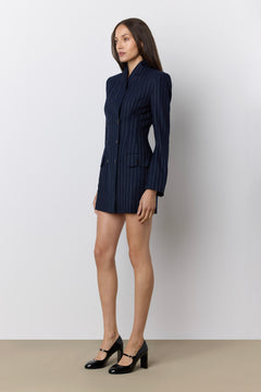 A woman modeling a navy blue pinstripe blazer dress with black Mary Jane heels, standing against a neutral light gray studio background.