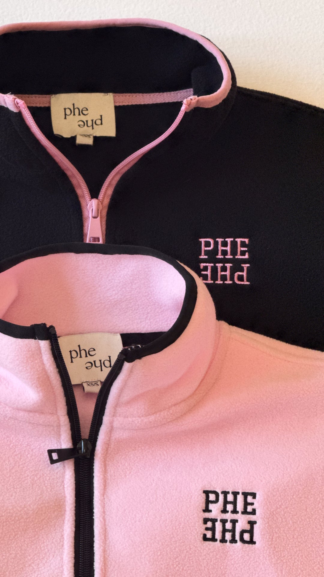 Two fleece quarter zip sweatshirts laying stacked on a table with embroidered "PHE PHE" lettering on left chest.