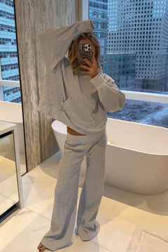 A woman takes a mirror selfie in a modern bathroom, wearing a light gray two-piece waffle knit lounge set consisting of a pullover hoodie and matching wide-leg sweatpants. A white freestanding tub and large windows showing a city skyline are visible in the background.