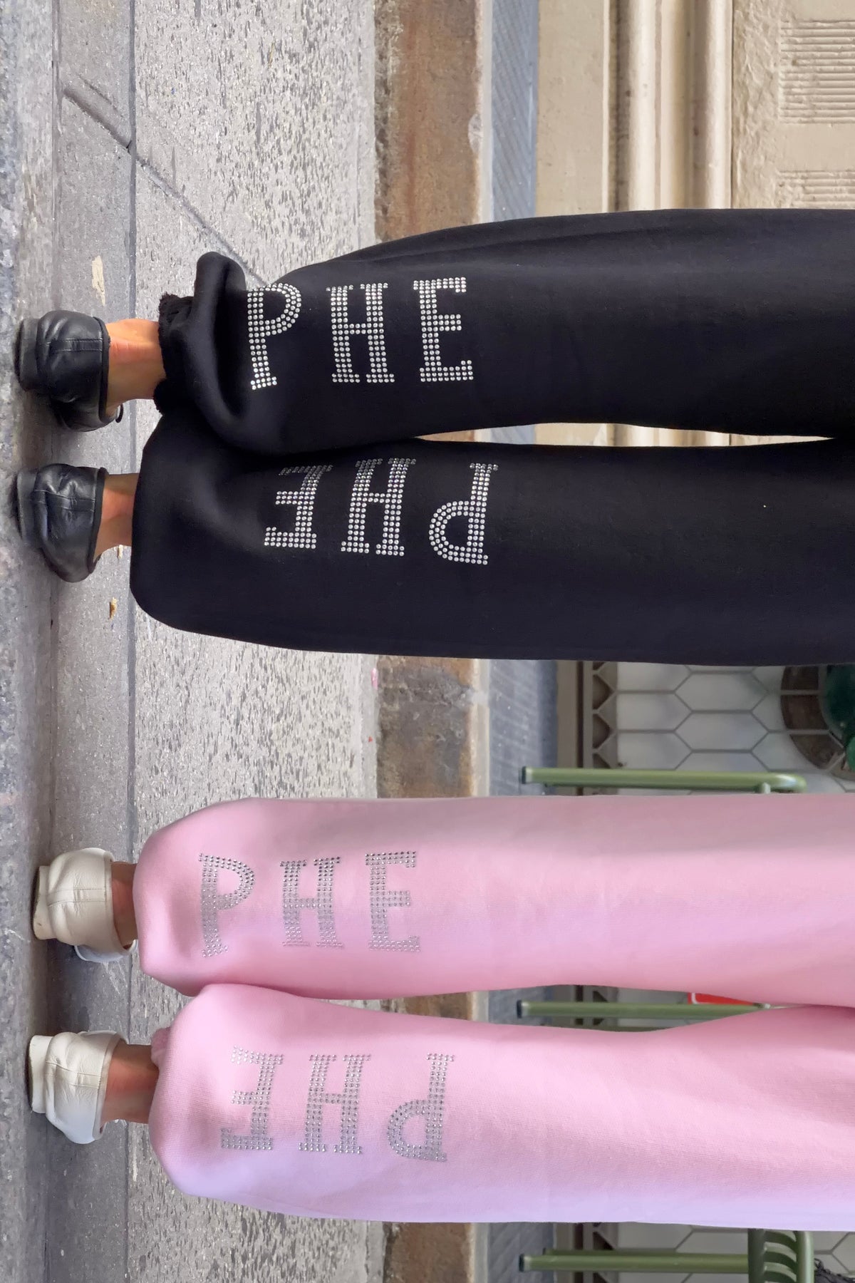 Two pairs of wide-leg sweatpants shown from the back, one black and one pink, both featuring the word 'PHE' in large silver rhinestone letters down the left leg, worn by two models standing on a sidewalk.