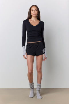 A woman stands against a light gray studio wall wearing a black long-sleeve top, black athletic shorts, and gray socks.