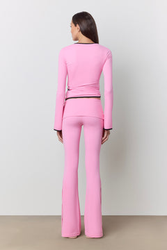 A model viewed from the back wearing a bright pink two-piece matching set consisting of a long-sleeve wrap-style top and flared pants, with thin black trim details at the collar, cuffs, and waist.