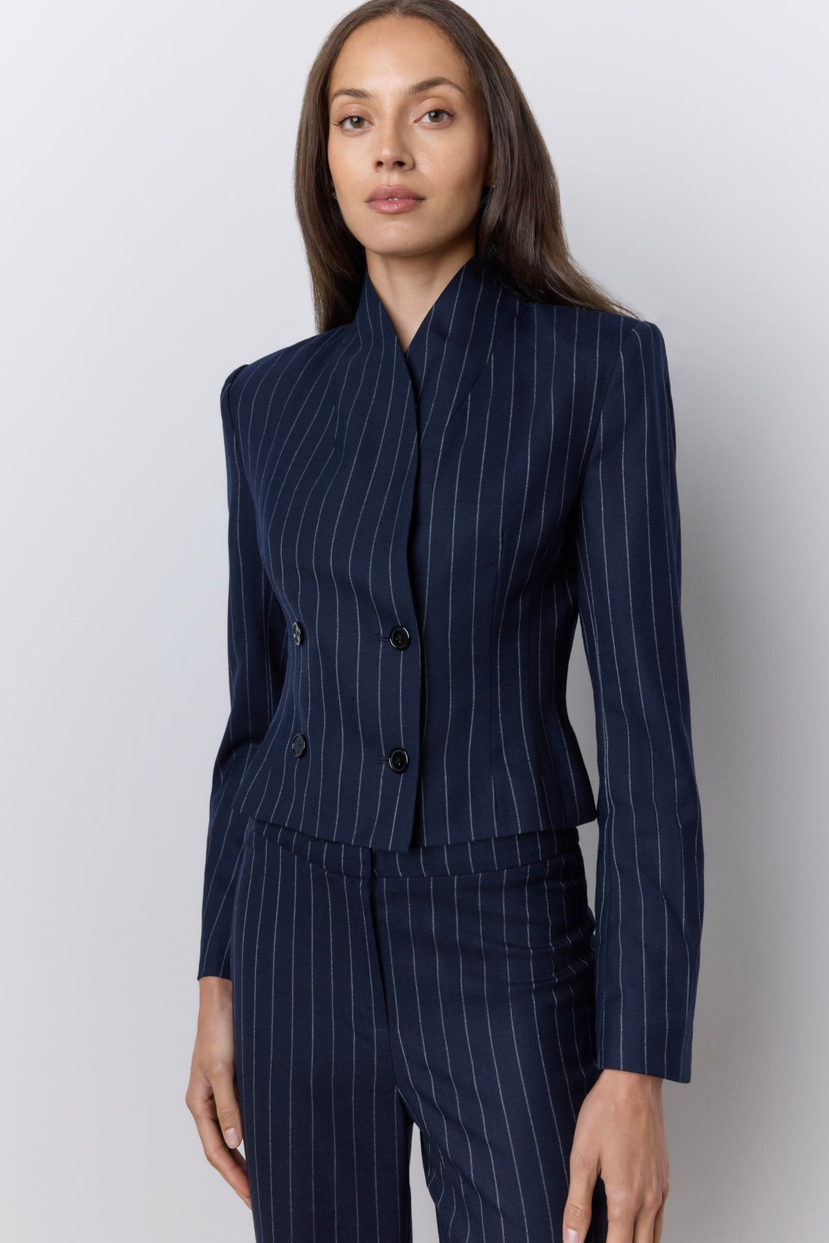A woman modeling a navy blue pinstripe double-breasted blazer with matching trousers against a light gray studio background.