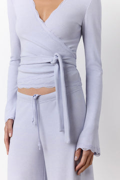 A close-up of a model wearing a light periwinkle wrap top with a tie waist and matching drawstring pants against a plain light background.