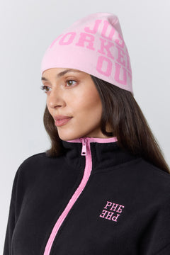 A woman wearing a light pink knit beanie with pink text reading 'JUST WORKED OUT' and a black fleece zip-up jacket with pink trim and matching pink text on the chest, photographed against a light gray studio background.