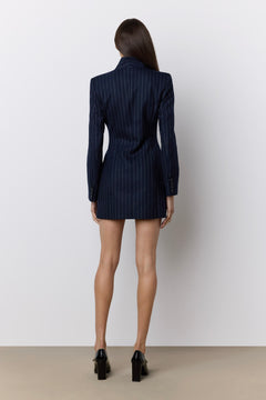 A woman stands with her back to the camera wearing a navy blue pinstripe blazer dress with long sleeves and black high heels, set against a plain light gray wall and beige floor.