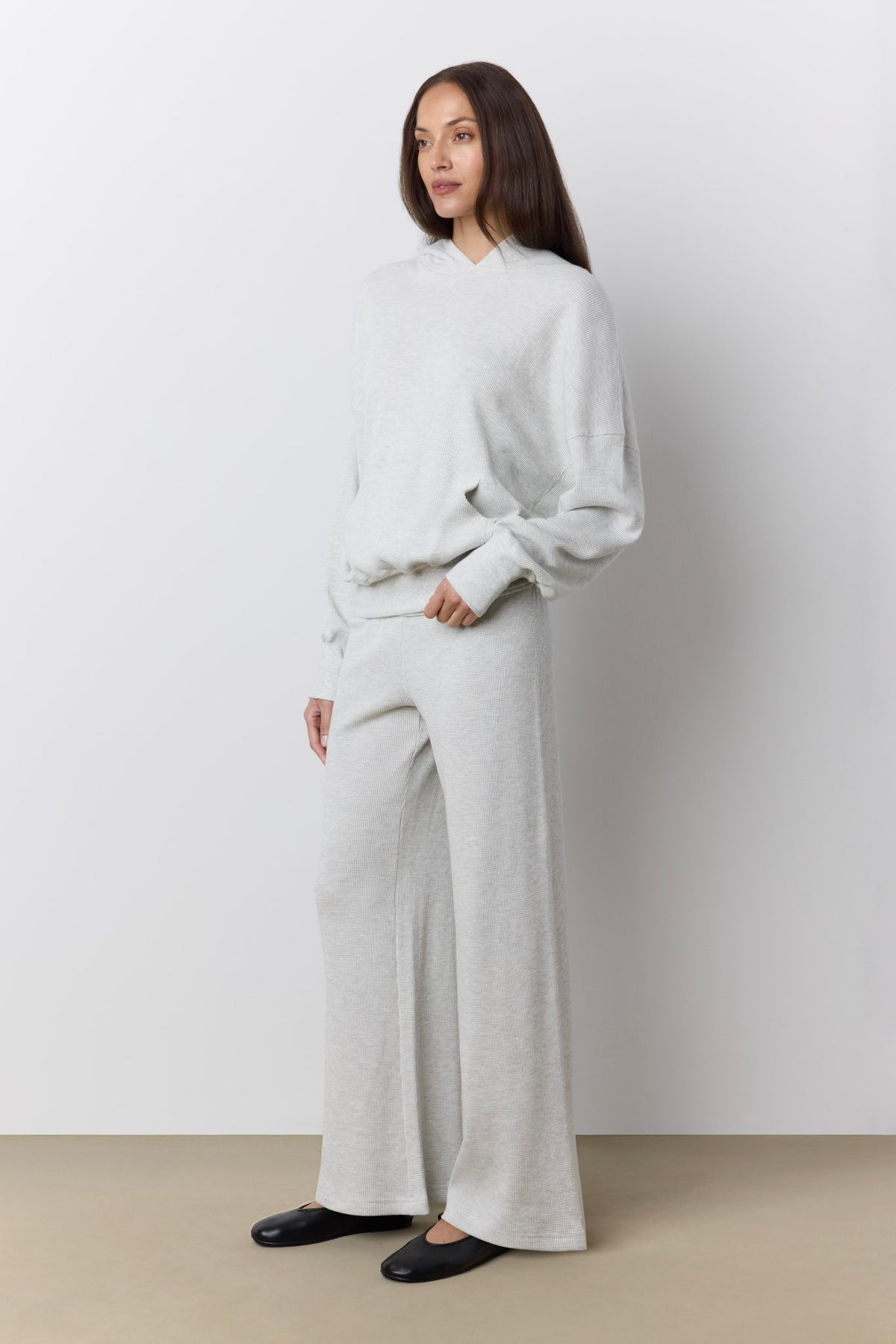 A woman modeling a light gray waffle knit lounge set consisting of a long-sleeve hoodie and matching wide-leg pants, paired with black flats, against a neutral studio backdrop.