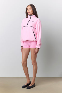 A woman modeling a light pink fleece quarter-zip sweatshirt and matching shorts, with black zipper accents and a small black embroidered "PHE PHE" logo on the chest, set against a neutral studio background.
