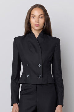 A model stands against a light gray studio wall wearing a tailored black two-piece suit with a structured double-breasted jacket and matching wide-leg trousers.