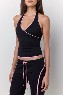 A woman modeling a black athletic halter crop top and matching black pants with bright pink trim and drawstring details, photographed against a light gray studio background.