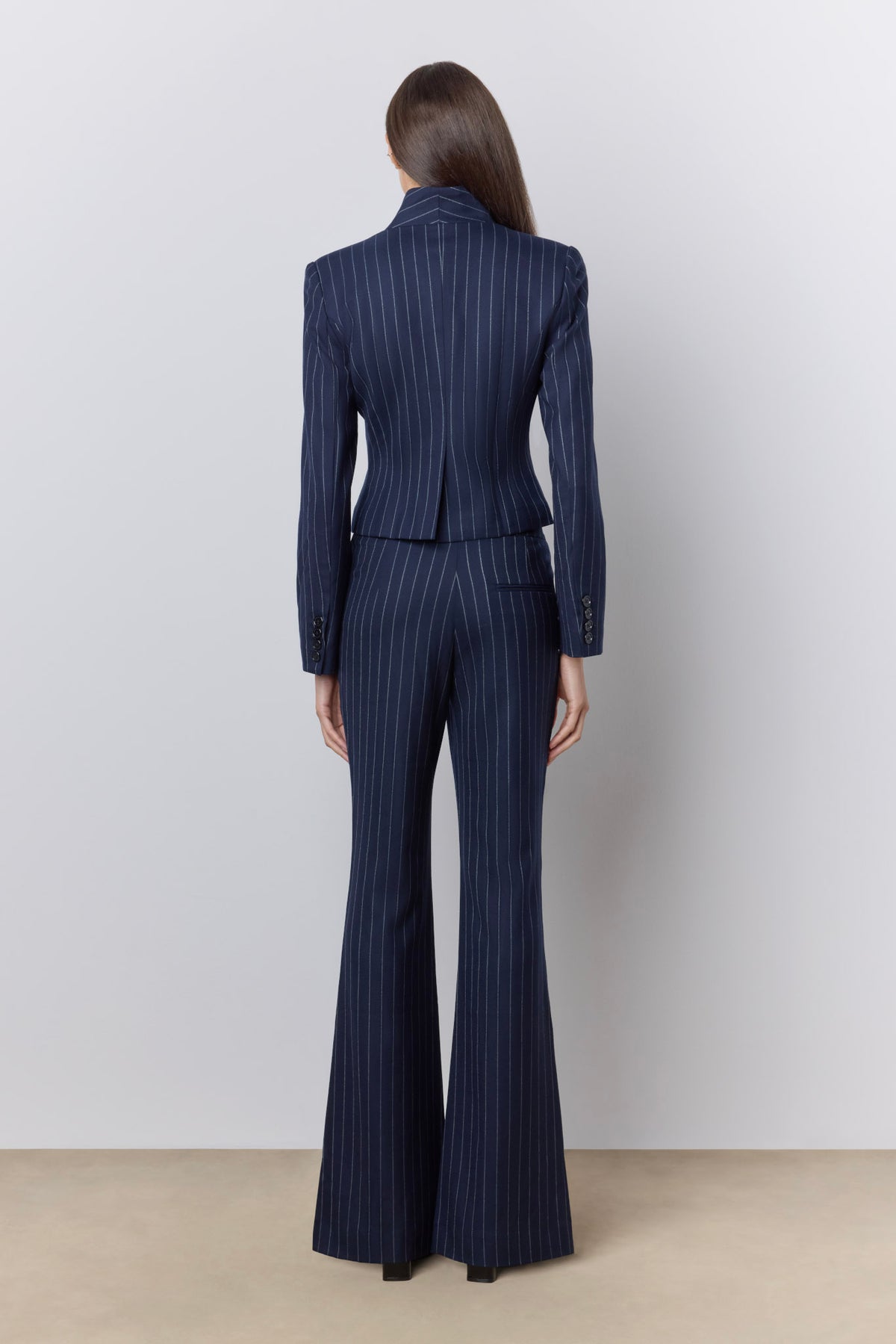 A model viewed from the back wearing a navy blue pinstripe blazer and matching flared trousers against a light gray studio background.