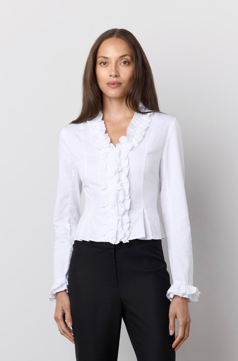 A woman modeling a white long-sleeve blouse with ruffle trim and black flared pants against a plain light gray studio background.