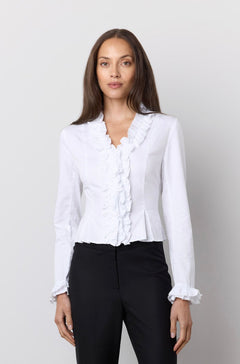 A woman modeling a white long-sleeve blouse with ruffle trim and black flared pants against a plain light gray studio background.