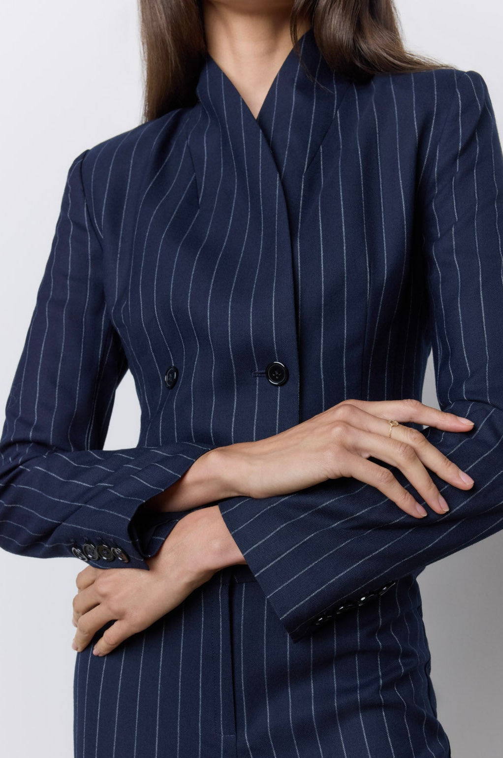 A woman wearing a navy blue pinstripe double-breasted blazer and matching trousers, shown from the neck down against a light gray studio background.