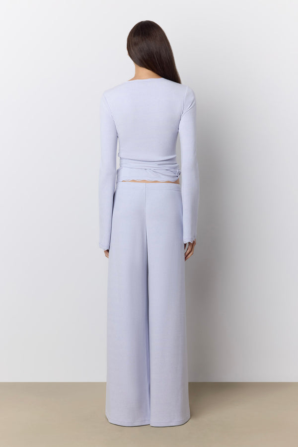 A model viewed from the back wearing a periwinkle two-piece set consisting of a long-sleeve wrap-style top and matching wide-leg lounge pants, set against a neutral studio background. |M1