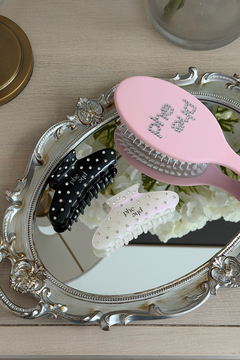 Two claw hair clips with rhinestones and the text "phe phe" are arranged on an ornate silver mirror, alongside a pink hairbrush with "phe phe" rhinestone lettering on a light wood tabletop.