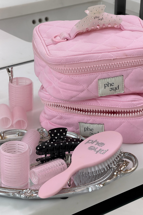 A pink quilted cosmetic bag with matching hair accessories, including a hairbrush, claw clips, and hair curlers, arranged on a silver tray. The brand name 'phe phe' is visible on the labels and hair accessories.