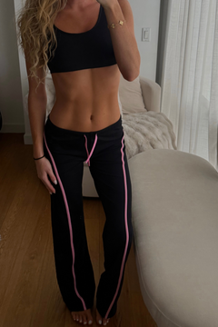A woman wearing a black sports bra and black wide-leg track pants with pink side stripes, standing in a home setting with a beige sofa and wood floor.