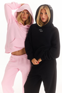 Two women modeling matching lounge sets against a white studio background. One wears a light pink hoodie and sweatpants, and the other wears a black hoodie with rhinestone "phe phe" logo on the chest and black sweatpants.