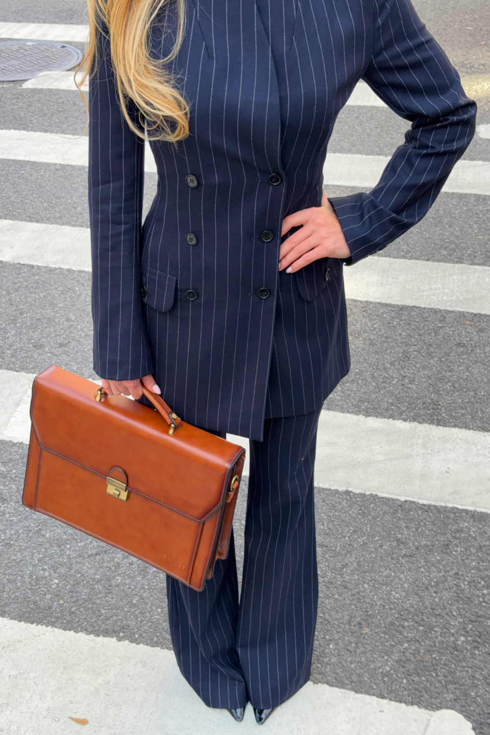 A person wearing a navy blue pinstripe double-breasted dress with matching trousers and holding a brown leather briefcase, standing on a street crosswalk.