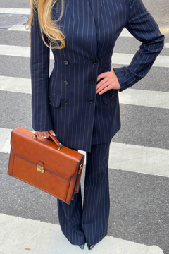 A person wearing a navy blue pinstripe double-breasted dress with matching trousers and holding a brown leather briefcase, standing on a street crosswalk.
